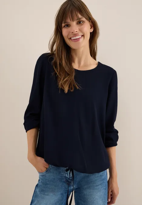 Frau mit dunklem Langarmshirt und blauer Jeans lächelt.